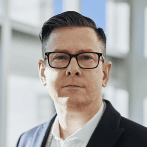 Geschäftsmann mit Brille blickt ernst in die Kamera, im Hintergrund ein Büro mit großen Fenstern.
