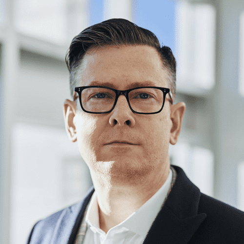 Geschäftsmann mit Brille blickt ernst in die Kamera, im Hintergrund ein Büro mit großen Fenstern.